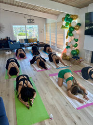 Bright living-room group yoga session with participants on colorful mats in child's pose, water bottles nearby, and a festive green-white-gold balloon column by sunlit sliding doors showing a city view.