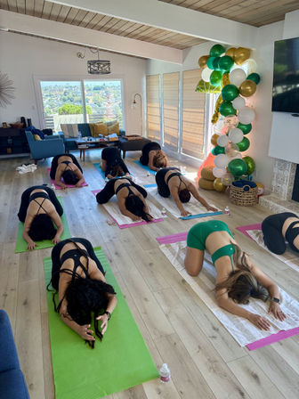 Bright living-room group yoga session with participants on colorful mats in child's pose, water bottles nearby, and a festive green-white-gold balloon column by sunlit sliding doors showing a city view.