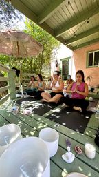 Sunlit outdoor yoga session on a green covered wooden deck — women meditating cross-legged on mats with an umbrella, string lights, crystal bowls, candle and water bottles amid leafy surroundings.