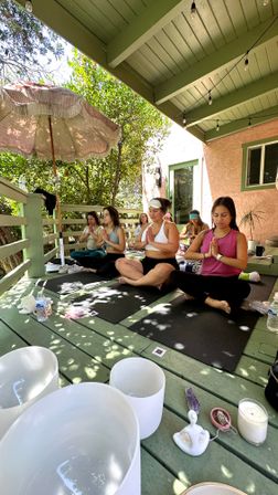 Sunlit outdoor yoga session on a green covered wooden deck — women meditating cross-legged on mats with an umbrella, string lights, crystal bowls, candle and water bottles amid leafy surroundings.