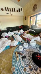 Cozy group sound bath in a sunlit living room — participants resting on yoga mats under white blankets, crystal singing bowls and healing instruments arranged on a blue mandala cloth, wooden carved figures on the high wall and sliding glass doors to a patio.