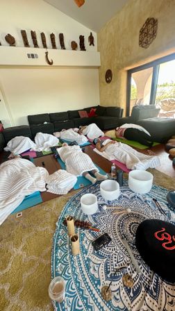 Cozy group sound bath in a sunlit living room — participants resting on yoga mats under white blankets, crystal singing bowls and healing instruments arranged on a blue mandala cloth, wooden carved figures on the high wall and sliding glass doors to a patio.