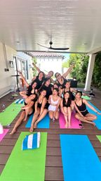 Group of women in activewear on colorful yoga mats on a covered backyard patio, smiling and posing after an outdoor yoga class.