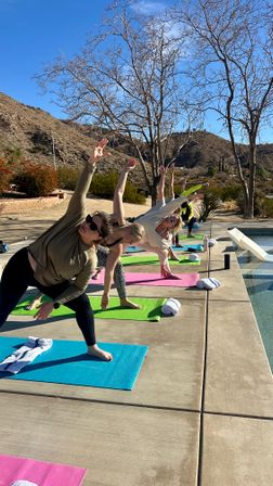 Poolside outdoor yoga class on colorful mats, participants stretching into side-angle poses by a swimming pool with bare trees and sunlit desert hills under a clear blue sky.