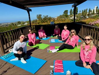 Seven women in matching pink "San Diego" shirts sitting on colorful yoga mats under a wooden pergola, smiling during an outdoor yoga/fitness session with coastal hills and an ocean view on a sunny day.