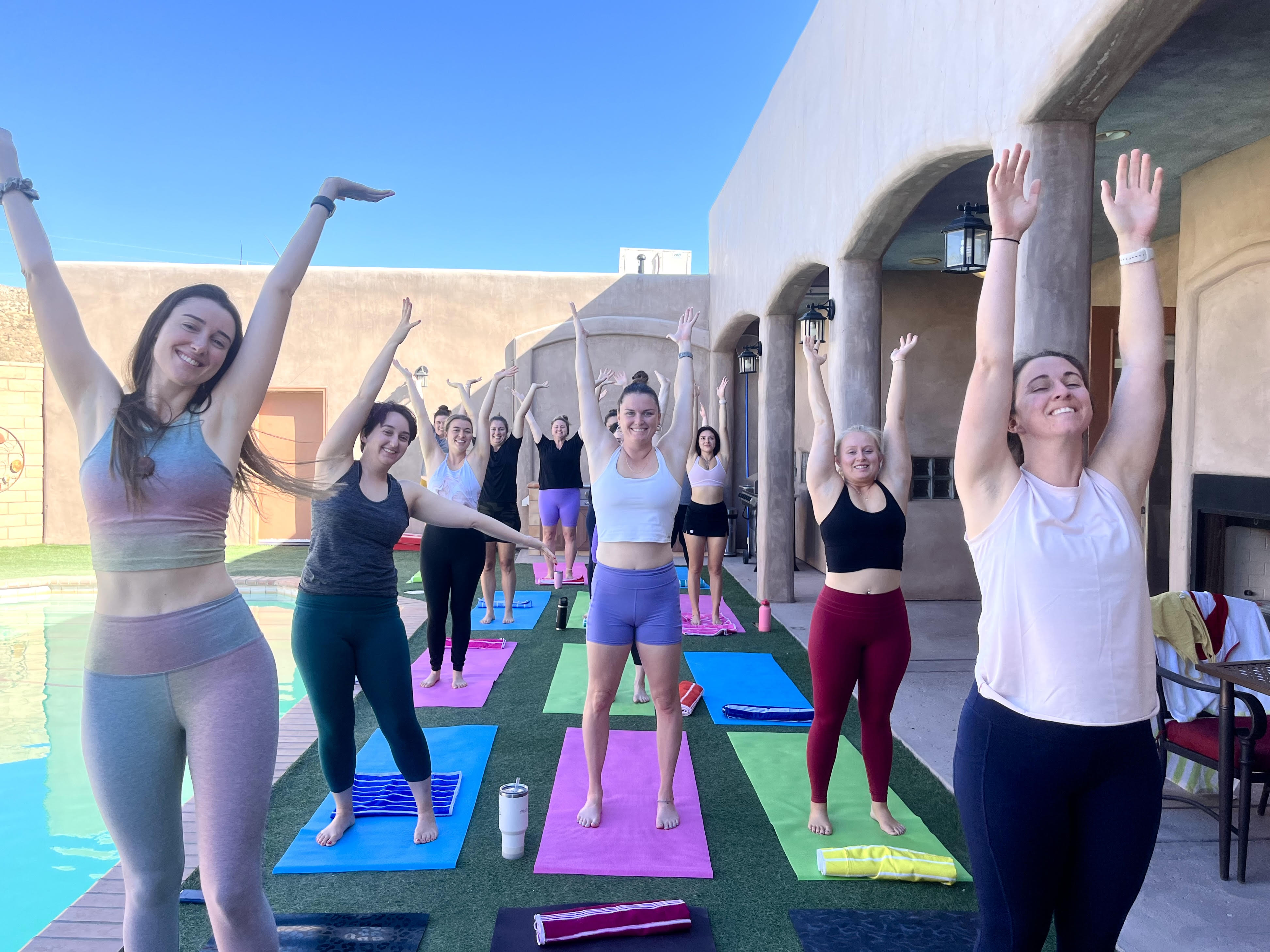 Smiling group of women doing a poolside outdoor yoga class on colorful mats, arms raised in a standing stretch on a sunny Southwestern-style patio with arches and bright blue sky.