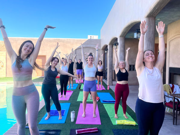 Smiling group of women doing a poolside outdoor yoga class on colorful mats, arms raised in a standing stretch on a sunny Southwestern-style patio with arches and bright blue sky.