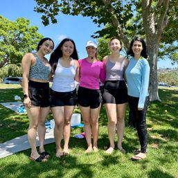 Five women in activewear smiling and posing barefoot on grass under trees at a sunny outdoor yoga/fitness class in a waterfront park