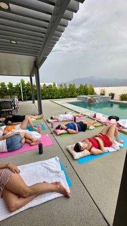 Poolside outdoor meditation session with several people lying on colorful yoga mats under a covered patio, sound bowls nearby and mountains in the background