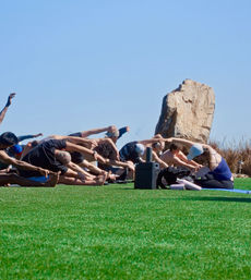 Sunny outdoor yoga class stretching in seated forward bends on a bright green lawn beside a large rock, with mats, water bottles, and a portable speaker visible.