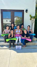 Seven women with colorful yoga mats seated on sunny suburban front steps in prayer pose in front of wooden double doors, lightbox sign reading HAPPY BACH DEMI!