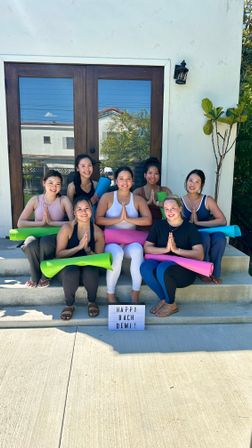 Seven women with colorful yoga mats seated on sunny suburban front steps in prayer pose in front of wooden double doors, lightbox sign reading HAPPY BACH DEMI!