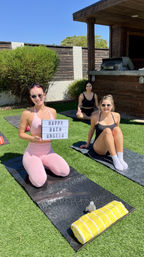 Three women in athletic wear on yoga mats in a sunny backyard lawn; front woman in pink holds a lightbox sign reading "HAPPY BACH ANGELA" for a bachelorette yoga party, yellow striped towel and water bottle on a mat, wooden patio and grill in background.