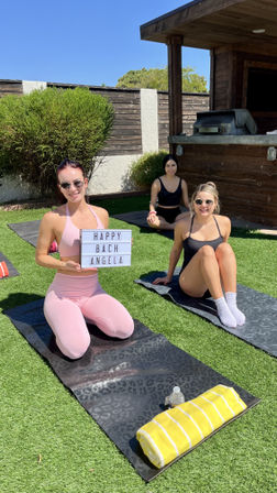 Three women in athletic wear on yoga mats in a sunny backyard lawn; front woman in pink holds a lightbox sign reading "HAPPY BACH ANGELA" for a bachelorette yoga party, yellow striped towel and water bottle on a mat, wooden patio and grill in background.