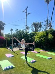 Person in sunglasses practicing dancer yoga pose on one leg in a sunny backyard with palm trees, green and pink mats and striped towels set up for an outdoor yoga class