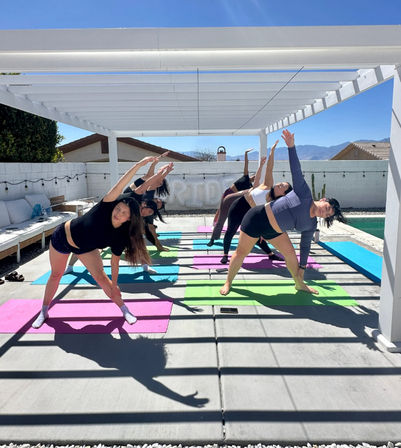 Sunny backyard yoga session: group of participants on colorful mats under a white pergola beside a pool, stretching into side-angle poses with mountains in the background.