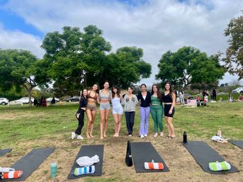 Eight women in activewear posing on a grassy public park lawn before an outdoor yoga class, with yoga mats, towels and leafy trees under a partly cloudy sky.