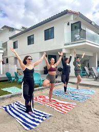 Four people balancing in tree pose on striped beach towels on sand in front of a modern beachfront house — group beach yoga session with Adirondack chairs and a glass balcony under a cloudy sky.