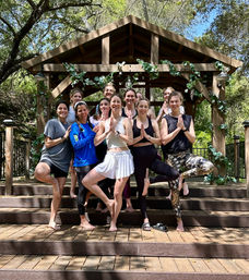 Smiling group of women in activewear practicing tree pose on wooden steps under a vine‑decorated gazebo in a sunlit wooded retreat