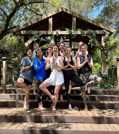 Smiling group of women in activewear practicing tree pose on wooden steps under a vine‑decorated gazebo in a sunlit wooded retreat