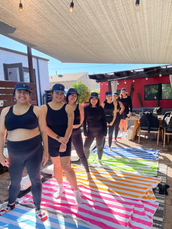 Smiling women in black activewear and matching caps lined up on bright striped towels under a fabric sunshade on a sunny backyard patio for a casual outdoor wellness meetup.