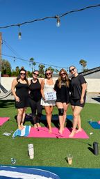 Five friends barefoot on pink yoga mats in a sunny backyard with palm trees and hanging string lights, smiling and holding a lightbox sign celebrating a bachelorette