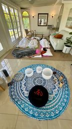 Sunlit sunroom sound-bath meditation with three people reclining on mats under blankets, crystal singing bowls and a blue mandala tapestry in the foreground