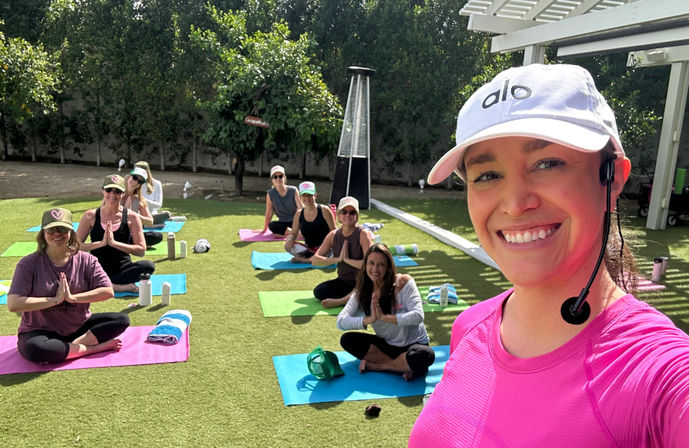 Smiling yoga instructor in a pink top and white cap with headset takes a selfie at a sunny outdoor yoga class on a green lawn, students on colorful mats seated in prayer pose near patio and trees.
