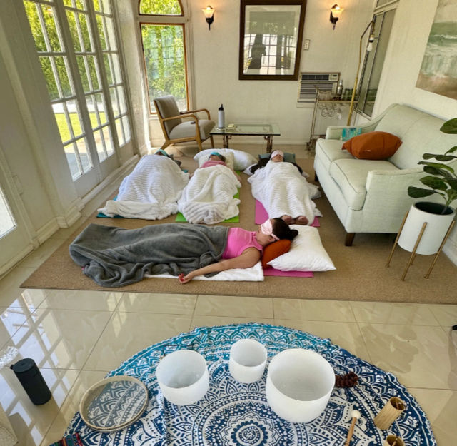 Sunlit living room group meditation and sound-bath setup: four people lying on yoga mats under blankets with eye masks, crystal singing bowls on a patterned mat in the foreground, glass doors, sofa and houseplants.