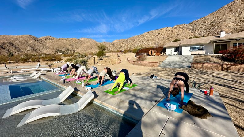 Poolside outdoor yoga class with people in downward dog on colorful mats beside a modern pool at a sunlit desert mountain retreat under a clear blue sky.