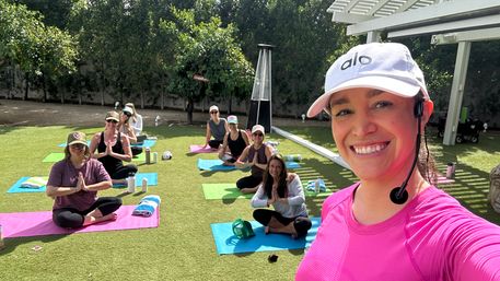 Outdoor yoga class on a sunny backyard lawn — smiling instructor in a pink shirt and white cap with headset takes a selfie while participants sit on colorful mats in prayer pose among trees.