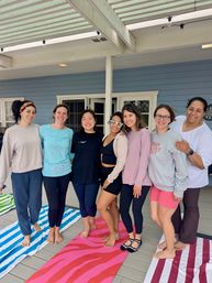 Seven women smiling and posing barefoot on colorful yoga mats on a covered outdoor deck with blue siding — casual group yoga or wellness gathering on a porch.