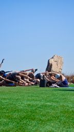 Coastal outdoor yoga class stretching on a vibrant green lawn beside a large rock under a bright blue sky