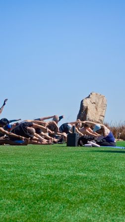 Coastal outdoor yoga class stretching on a vibrant green lawn beside a large rock under a bright blue sky