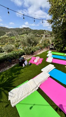 Outdoor yoga and sound bath setup on a grassy terrace with colorful pink, blue and green mats, rolled white blankets, crystal singing bowls on a pink cloth, speaker and string lights overlooking scenic green hills and valley under a sunny sky.