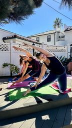 Two women in activewear doing side-stretch yoga on colorful mats on a sunny backyard deck with a white lattice fence and palm tree
