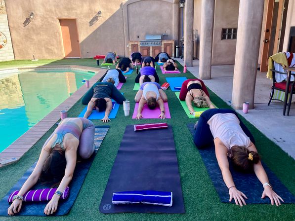 Outdoor poolside yoga class on a backyard patio: rows of participants in child’s pose on colorful mats on artificial turf beside a swimming pool, with water bottles, towels and a grill in the background.
