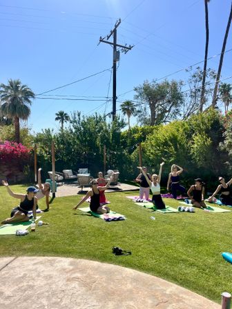 Outdoor yoga class on a sunny backyard lawn with palm trees, string lights and patio seating; participants on colorful mats stretching and waving under a clear blue sky.