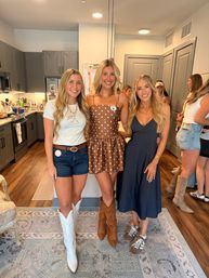 Three smiling women posing in a modern apartment kitchen — white cowboy boots with denim shorts, brown polka-dot dress with tan boots, and a navy midi dress with sneakers; hardwood floors, area rug and guests in the background.