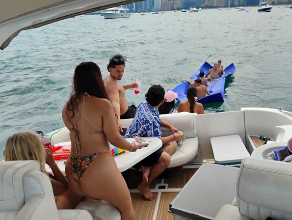 Friends partying on a yacht near a city skyline, lounging with drinks while others float on blue foam mats in choppy lake water.