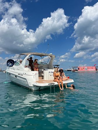 Friends lounging on a white motorboat in turquoise water under a bright blue sky with puffy clouds, summer boating scene
