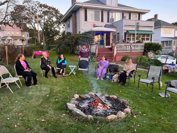 Friends gathered around a glowing stone fire pit on a suburban backyard lawn at dusk, folding chairs and a lit porch and deck of a two-story house in the background.