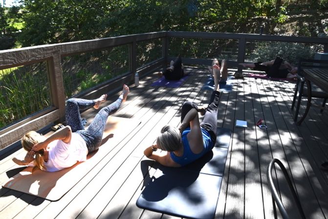 Sun-dappled outdoor fitness class on a wooden deck with participants on yoga mats doing core/bicycle-crunch exercises surrounded by trees.