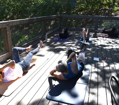Sun-dappled outdoor fitness class on a wooden deck with participants on yoga mats doing core/bicycle-crunch exercises surrounded by trees.