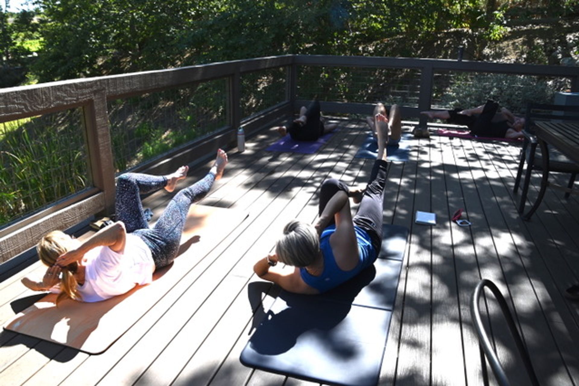 Sun-dappled outdoor fitness class on a wooden deck with participants on yoga mats doing core/bicycle-crunch exercises surrounded by trees.