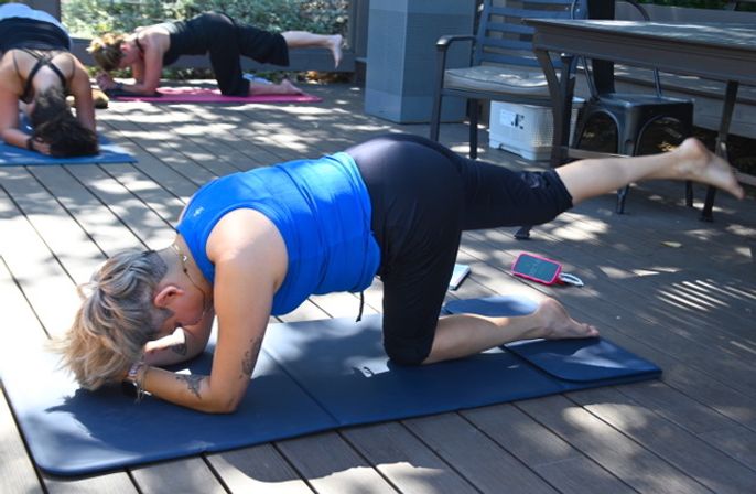 Person in a blue tank top doing a bird-dog yoga pose (forearm with one leg raised) on a mat during an outdoor group class on a sunlit wooden deck patio.