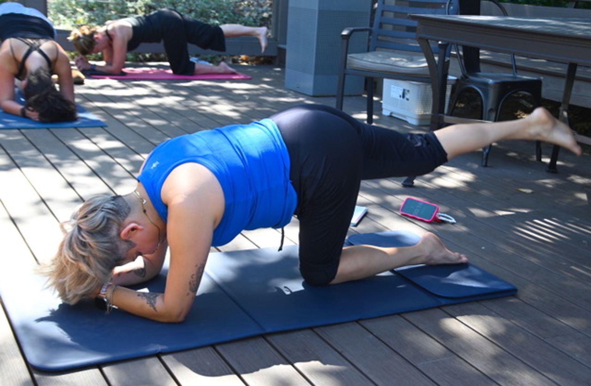 Person in a blue tank top doing a bird-dog yoga pose (forearm with one leg raised) on a mat during an outdoor group class on a sunlit wooden deck patio.