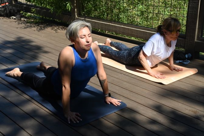 Two people practicing upward-facing dog yoga on mats on a sunny wooden deck with green foliage behind a railing.