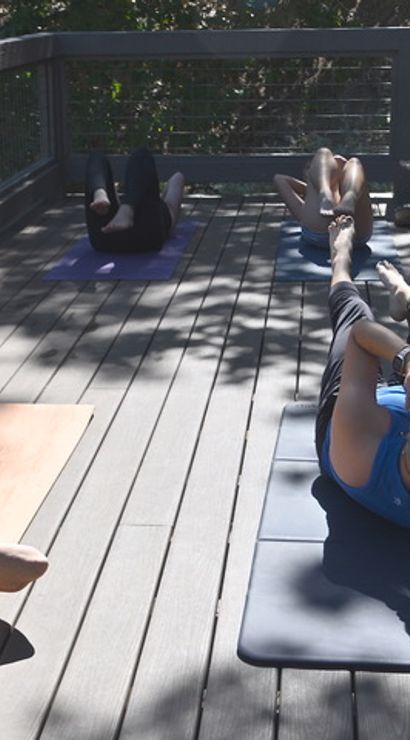 Outdoor core workout with participants doing bicycle crunches on yoga mats on a sunny wooden deck surrounded by trees