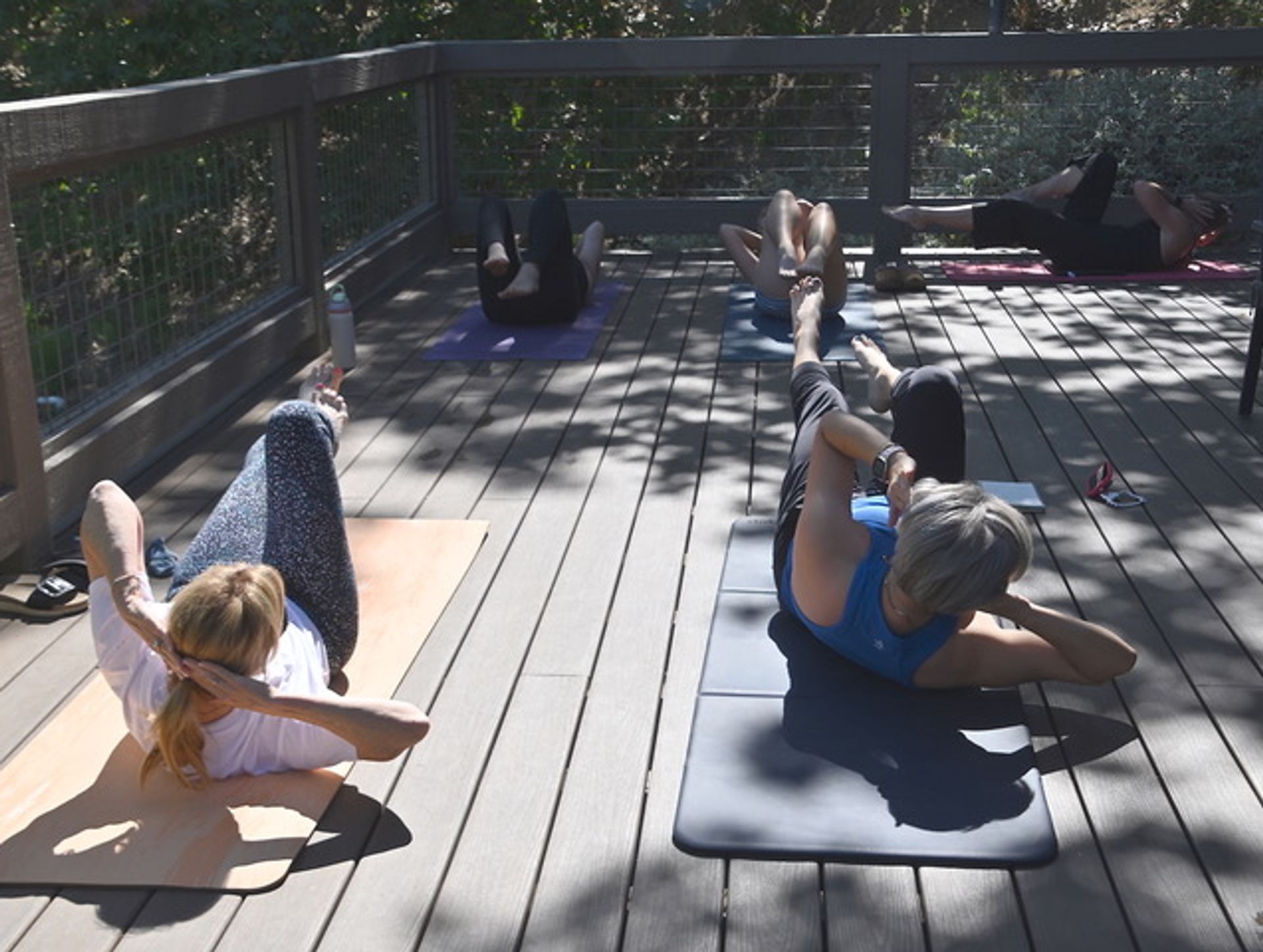 Outdoor core workout with participants doing bicycle crunches on yoga mats on a sunny wooden deck surrounded by trees